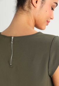 Olive green top with short sleeves, featuring a back zipper closure and textured fabric. Sleek, smooth finish with a subtle sheen.