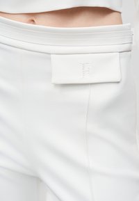 Κοντινό πλάνο στη μέση με λευκό παντελόνι ραμμένο στα μέτρα, με ανάγλυφο κεντητό γράμμα "F" στη θήκη της μπροστινής τσέπης.