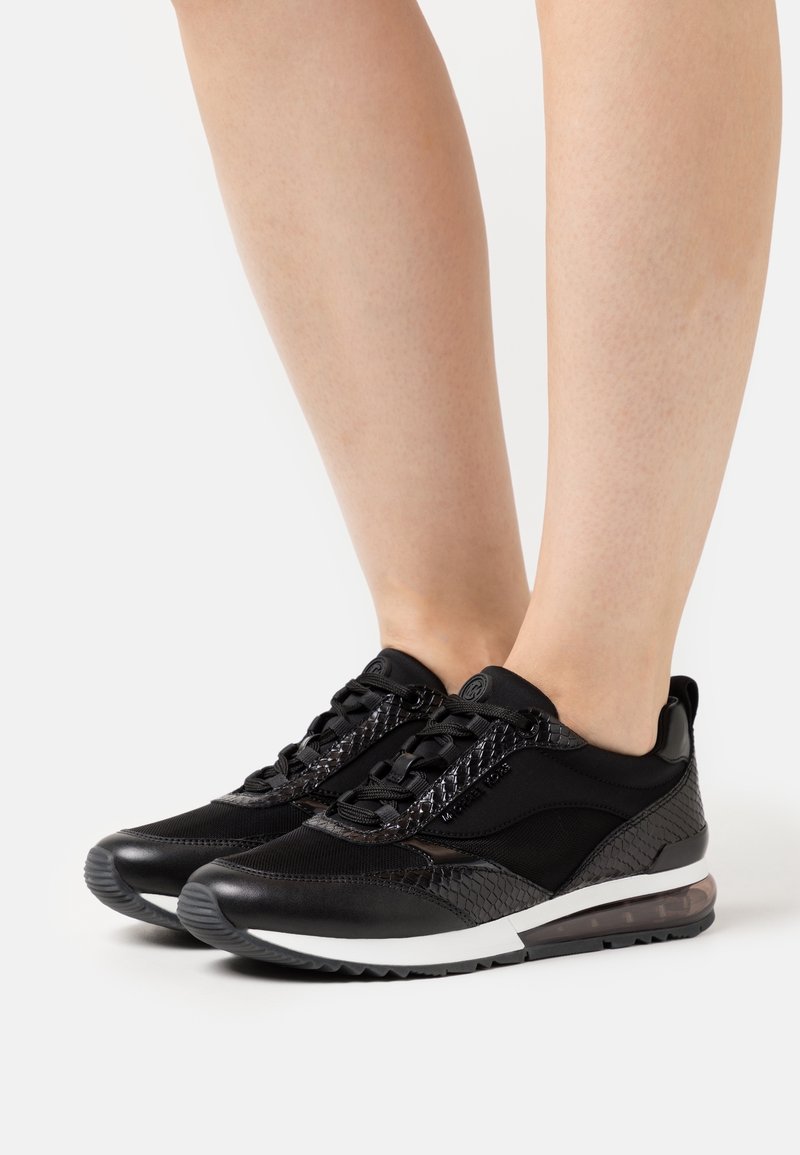 Paire de baskets noires avec des accents texturés portées pieds nus sur un fond blanc uni.
