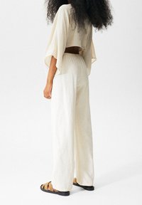 Beige crop top med brede ærmer og åben ryg, parret med løstsiddende cremefarvede bukser og brune sandaler med stropper. Blød tekstur.