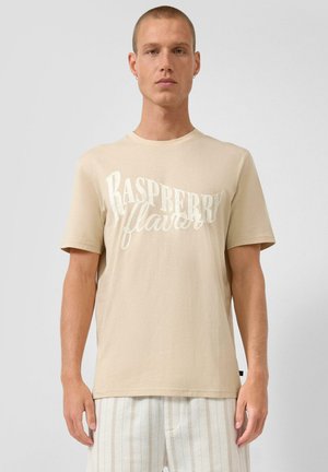 Jeune homme portant un T-shirt beige avec le texte "Saveur Framboise" et un pantalon léger rayé, debout devant un fond clair uni.