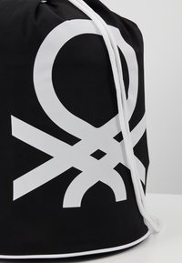 Sac à dos à cordon noir fabriqué en matériau durable, avec de grands motifs graphiques blancs, comprenant des cercles et des lignes entrecroisées, accompagné d'un cordon blanc.