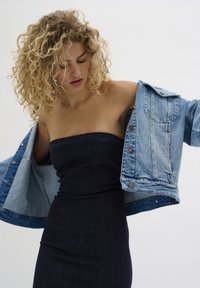 Una giacca di denim azzurro chiaro è indossata sopra un abito senza spalline di denim scuro. La giacca presenta bottoni metallici e una texture leggermente consumata.