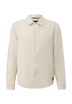 Beige Hemdjacke mit langen Ärmeln, Kragen, Druckknopf-Verschluss vorne und einem kleinen schwarzen Etikett nahe dem Saum.