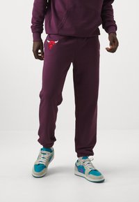 Pantaloni della tuta viola con logo del toro rosso ricamato e polsini a costine, abbinati a scarpe da ginnastica multicolori con accenti blu e viola.