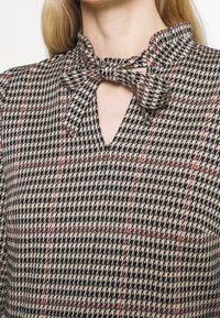 Blouse à carreaux noir et beige avec un col froncé, décolleté en trou de serrure et un détail de nœud lié. Présente des lignes rouges accentuant le motif.
