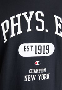 Černé bavlněné tričko s výrazným bílým nápisem "PHYS. ED" a "EST. 1919." a malým červeným a bílým logem, navržené značkou Champion, New York.
