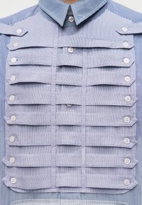 Κοντινό πλάνο μπλε πουκαμίσου με ρίγες, με επάλληλα οριζόντια υφασμάτινα πάνελ που στερεώνονται με λευκά κουμπιά στην περιοχή του στήθους.