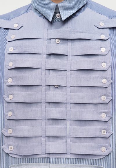 Gros plan sur une chemise à fines rayures bleues avec des rabats horizontaux superposés maintenus par des boutons blancs au niveau de la poitrine.