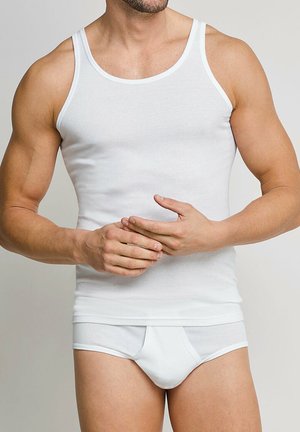 Mann trägt eng anliegendes weißes ärmelloses Unterhemd und passende Slips, steht vor einfarbigem Hintergrund mit den Händen vor der Taille verschränkt.