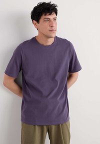 Fialové bavlněné tričko s kulatým výstřihem a krátkými rukávy, v kombinaci s texturovanými olivově zelenými kalhotami. Jednoduchý design, minimální detaily.