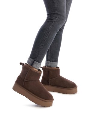 Botas de tobillo de ante marrón con una textura suave, puntera redonda, suela plana de goma y una tira de agarre. Llevadas con vaqueros oscuros doblados.