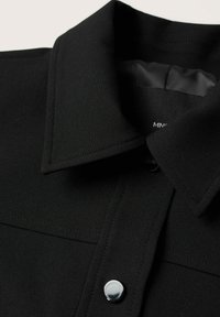 Chemise noire à col en tissu lisse avec une finition mate, dotée d'un seul bouton-pression brillant et d'un motif discret cousu sur le col.