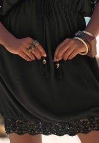 Robe noire en tissu texturé, avec un ourlet en dentelle et des accents à franges. Mains ornées de bagues décoratives et de multiples bracelets.