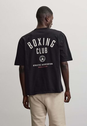 Hombre de espaldas a la cámara, vistiendo una camiseta negra con el texto "Boxing Club Athletic Association Pegador" y pantalones beige.