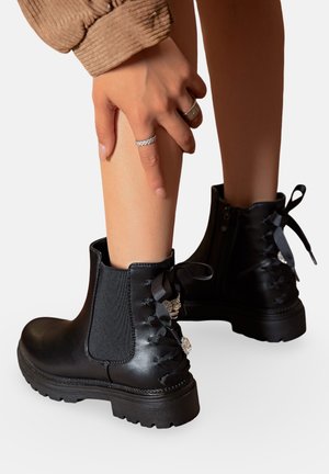 Bottines noires en cuir lisse avec des panneaux latéraux élastiques, un détail de laçage à l'arrière et une quincaillerie décorative. Semelle en caoutchouc épaisse.