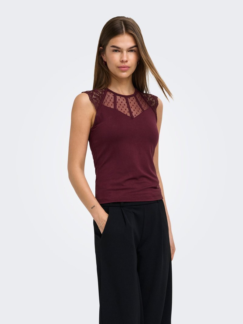 Jeune femme aux cheveux bruns raides porte un haut bordeaux sans manches avec des détails en dentelle et en résille, associé à un pantalon noir à jambes larges.
