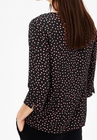 Blusa nera con pois rosa, maniche a tre quarti, spacchi laterali e una texture morbida. Presenta un collo rotondo e una vestibilità ampia.