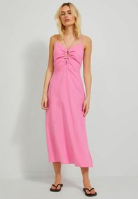 Vestido midi rosa con tirantes finos, parte delantera fruncida y detalle de ojal. Tejido ligero con una textura suave y una silueta fluida. Sandalias negras.