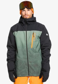 Chaqueta de esquí impermeable verde y negra con un bolsillo con cremallera y acentos naranjas. Se lleva con guantes negros y un gorro de punto naranja.