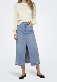 Mujer con un suéter de punto color crema, una falda midi de mezclilla azul clara con abertura frontal, bailarinas negras y un collar de cadena dorada.