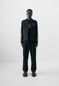 Veste en denim noir avec texte jaune, t-shirt noir en dessous et pantalon noir. Porté avec des chaussures noires. Design simple et ajustement.
