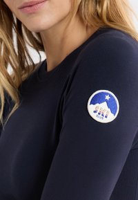 Granatowa bluzka z długim rękawem wykonana z gładkiego materiału, ozdobiona okrągłą naszywką z haftowanym wzorem góry i napisem "SKI CLUB".