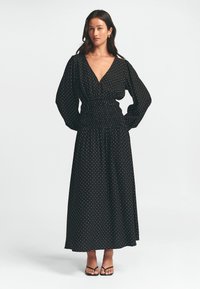 Černé maxi šaty s puntíky, hlubokým výstřihem do V, stahovaným pasem, dlouhými rukávy a splývavou sukní. Doplněné černými sandály na tenkých řemíncích.