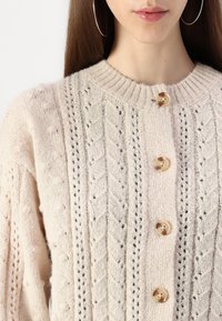 Cárdigan de punto color crema con un patrón texturizado, cuello acanalado y grandes botones de madera en la parte delantera.