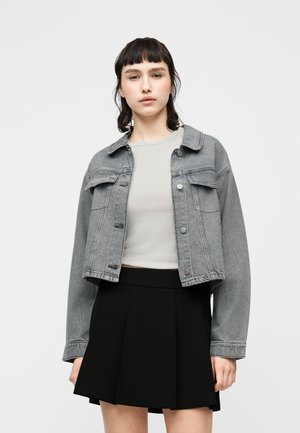 Mladá osoba s tmavými vlasy, oblečená v šedé krátké džínové bundě, bílém vršku a černé plisované sukni, stojící před jednoduchým pozadím.