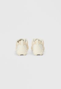 Adidași crem cu suprapunere albă texturată și tălpi cu țepi, având un inel de prindere în spate și un finisaj neted.