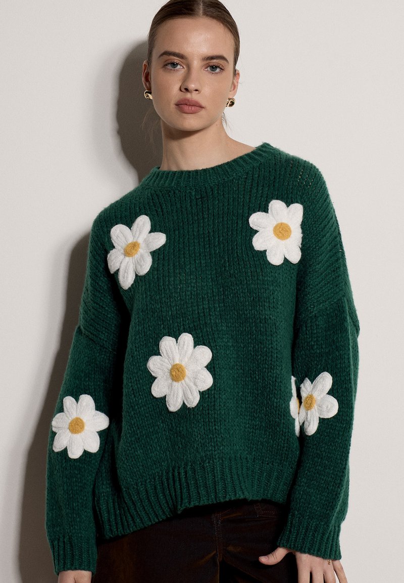 Femme portant un pull en tricot vert foncé avec de grandes marguerites blanches aux centres jaunes, debout devant un mur clair uni.