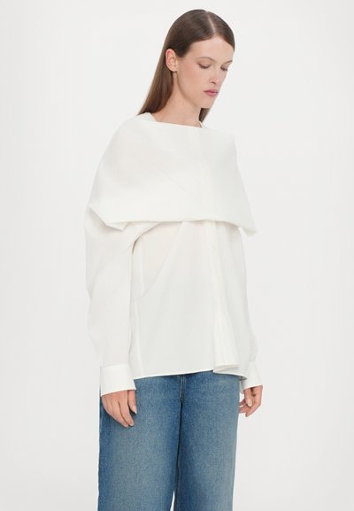 Witte oversized shirt met een unieke gedrapeerde bovenlaag, voorzien van een knoopsluiting aan de voorkant en lange mouwen, gecombineerd met brede blauwe denim broek.