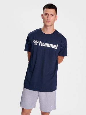 Mand står med hænderne på ryggen iført navyblå t-shirt med "hummel"-logo og lysegrå shorts mod en ensfarvet lys baggrund.