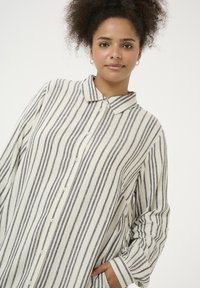 Camicia colletto con maniche lunghe, caratterizzata da strisce verticali crema e grigio scuro, chiusura con bottoni davanti e tasca sul lato destro.