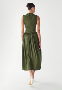 Maslinasto zelena haljina bez rukava s prilagođenim korzetom i naboranom suknjom, koja ima zatvaranje na munje sa stražnje strane. Glatka tkanina s mat završnom obradom.