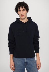 Felpa blu navy con un grande logo in rilievo, collo con cordino e tasca anteriore con polsini a coste. Tessuto morbido con una consistenza liscia.