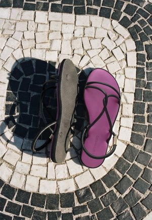 Paire de sandales violettes avec des brides noires sur un pavement en mosaïque circulaire noir et blanc, une semelle de sandale tournée vers le haut, projetant une ombre.