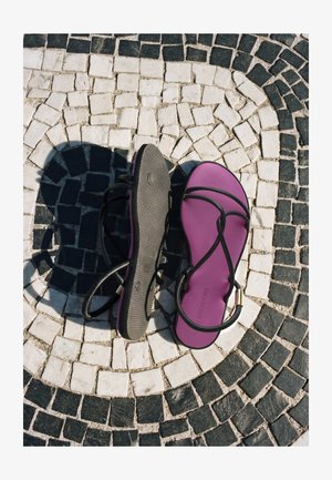 Paire de sandales violettes avec des brides noires sur un pavement en mosaïque circulaire noir et blanc, une semelle de sandale tournée vers le haut, projetant une ombre.