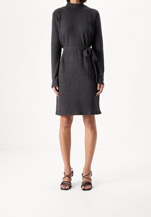 Kvinde iført en mørkegrå strikket kjole med lange ærmer og bælte i taljen, kombineret med sorte rem-sandaler med høj hæl, stående mod en hvid baggrund.