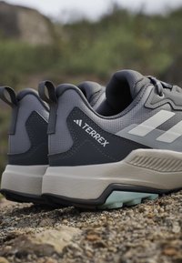 Paire de chaussures de trail Adidas Terrex grises posées sur un terrain rocheux avec un accent sur les talons et les semelles intermédiaires texturées.