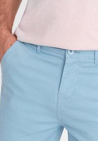 Světle modré bavlněné kalhoty s hladkou texturou, které mají přední kapsy a zapínání na knoflík. Detail šití je viditelný poblíž pásku.