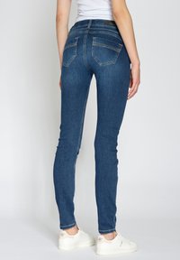 Úzké džíny v tmavě modré barvě, s dvěma zadními kapsami, jemnými švy a hladkou texturou. Stylizované s bílými teniskami.