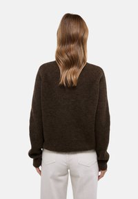 Femme aux cheveux bruns ondulés mi-longs portant un pull en tricot marron foncé et un pantalon beige clair, vue de dos.