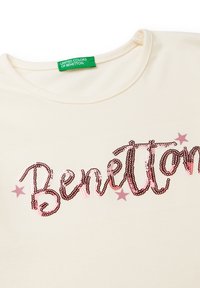 Kremowa koszulka z logo "Benetton" wyszywanym cekinami w odcieniach różu, z małymi różowymi gwiazdkami w pobliżu napisu.