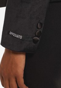 Chaqueta de traje negra con una textura suave, que presenta tres botones de satén negro y una etiqueta "OPPOSUITS" en el puño.