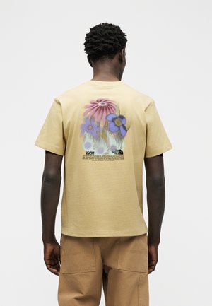 Mann som står med ryggen til, iført beige T-skjorte med blomstermotiv og tekst, kombinert med brune bukser, foran en ensfarget hvit bakgrunn.