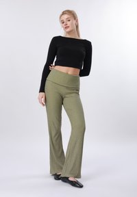 Νέα γυναίκα όρθια, φοράει μαύρο crop top με μακριά μανίκια, ελαιοπράσινο ριμπ παντελόνι και μαύρα επίπεδα παπούτσια, πάνω σε απλό φωτεινό φόντο.