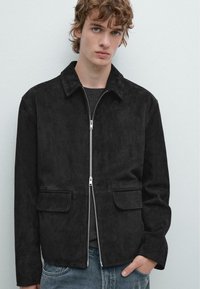 Schwarze Wildlederjacke mit einem Kragen, Frontreißverschluss und zwei aufgesetzten Taschen. Glatte Textur und strukturierte Passform, kombiniert mit lässigen Jeans.