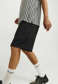 Osoba kráčející v černých šortkách, černých kožených botách, bílých značkových ponožkách a vodorovně pruhovaném černo-bílém tričku.